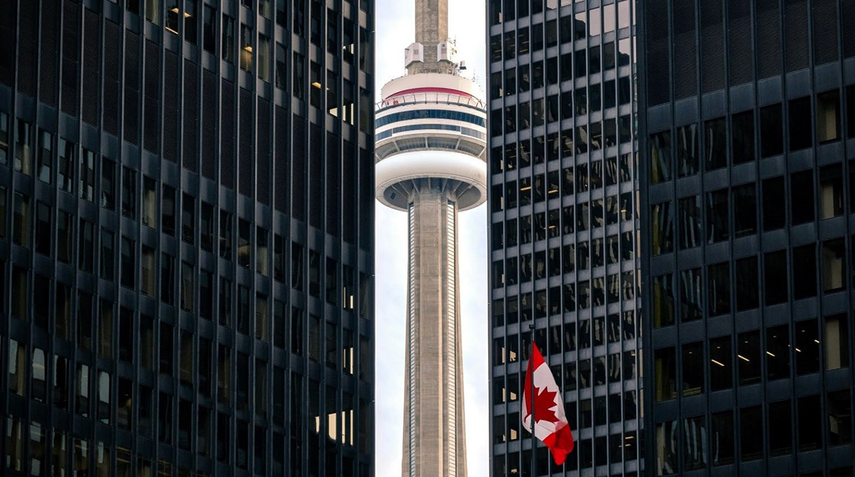 photo of CN tower between two highrises in Toronto with the Canadian flag flying in the foreground