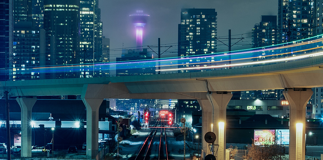 a trail of lights on an elevated roadway at night with commercial buildings in the background